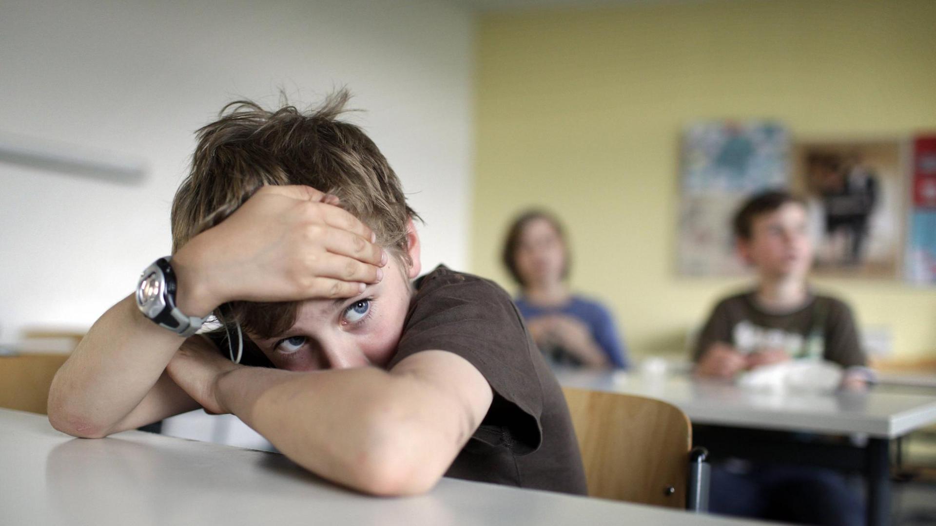 Ein Junge mit dunklem T-Shirt und zerzausten Haaren sitzt im Klassenzimmer und hat den Kopf auf die verschränkten Arme gestützt. Ein Junge mit dunklem T-Shirt und zerzausten Haaren sitzt im Klassenzimmer und hat den Kopf auf die verschränkten Arme gestützt.