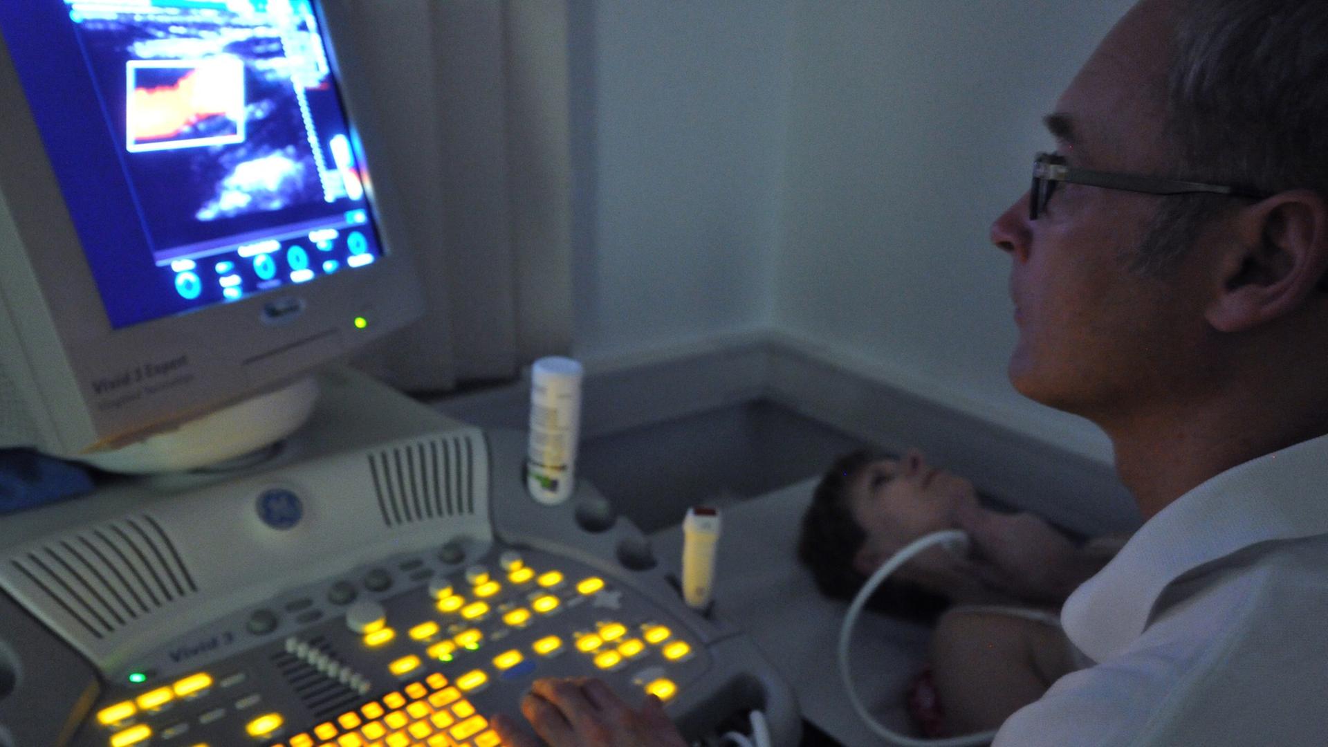 Ein Arzt sitzt in einem dunklen Raum vor einem Bildschirm eines Ultraschallgeräts, in der rechten Hand ein Ultraschallgerät, das an der Halsschlagader einer Patientin liegt. Ein Arzt sitzt in einem dunklen Raum vor einem Bildschirm eines Ultraschallgeräts, in der rechten Hand ein Ultraschallgerät, das an der Halsschlagader einer Patientin liegt.
