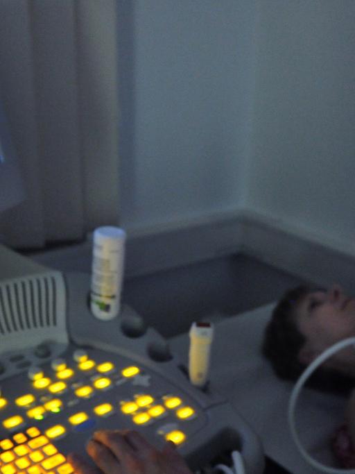 Ein Arzt sitzt in einem dunklen Raum vor einem Bildschirm eines Ultraschallgeräts, in der rechten Hand ein Ultraschallgerät, das an der Halsschlagader einer Patientin liegt.