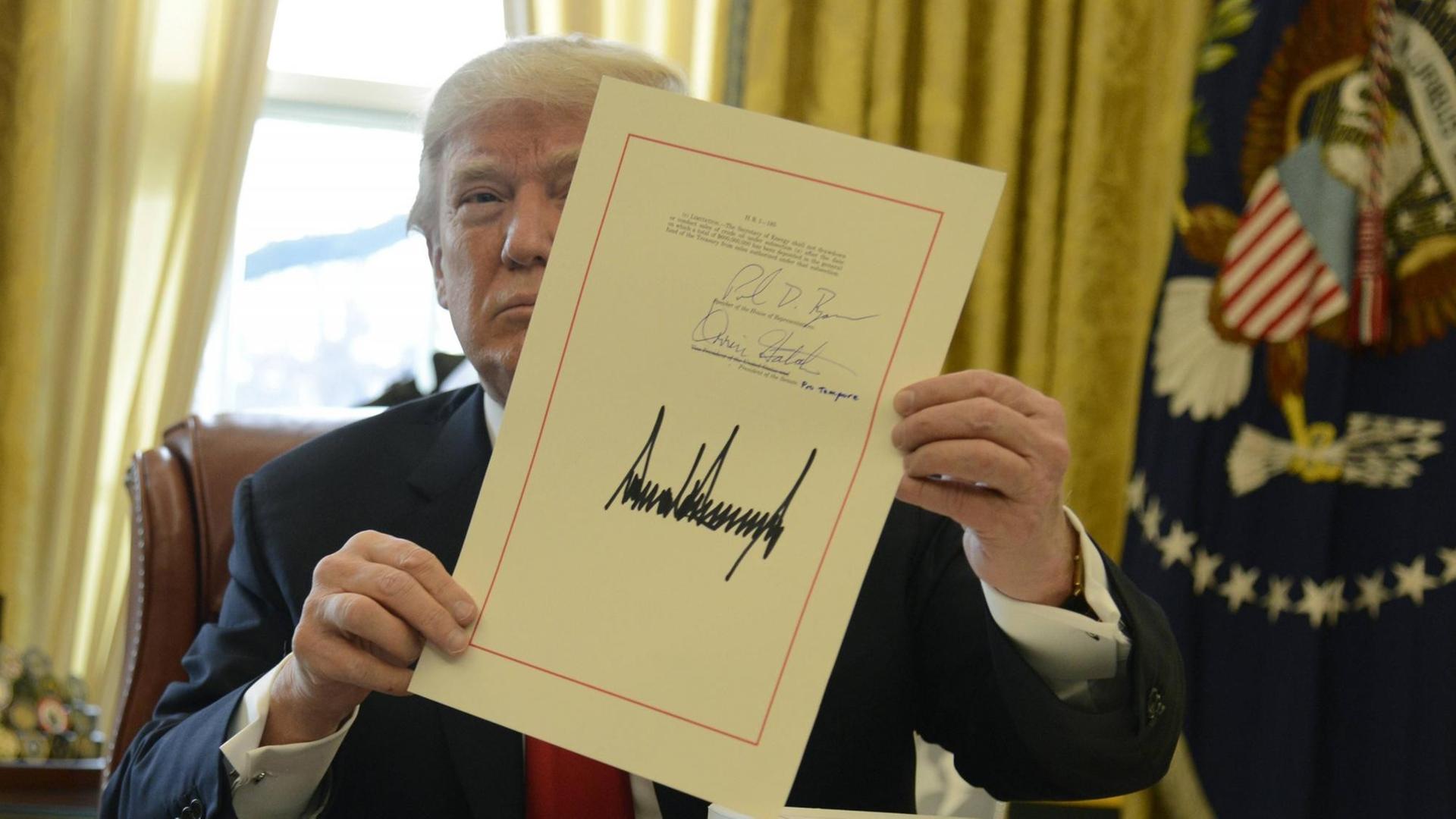 US-Präsident Donald Trump am 22. Dezember 2017 sitzt am Schreibtisch im Oval Officeim Weißen Haus und hält ein Dokument hoch, auf dem sehr groß seine Unterschrift zu sehen ist.