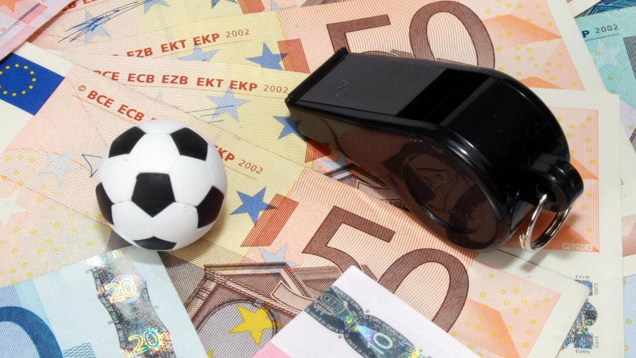 Auf einem Haufen Euro-Banknoten liegen ein Fußball und eine Schiedsrichterpfeife. Auf einem Haufen Euro-Banknoten liegen ein Fußball und eine Schiedsrichterpfeife.