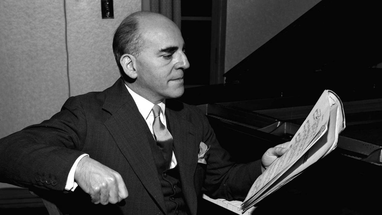 Der Pianist Solomon Cutner sitzt bei seinen morgendlichen Übungen am Klavier und hält Noten in der Hand. Er trägt einen Anzug mit Schlips. Schwarz-weiß Foto von 1953


