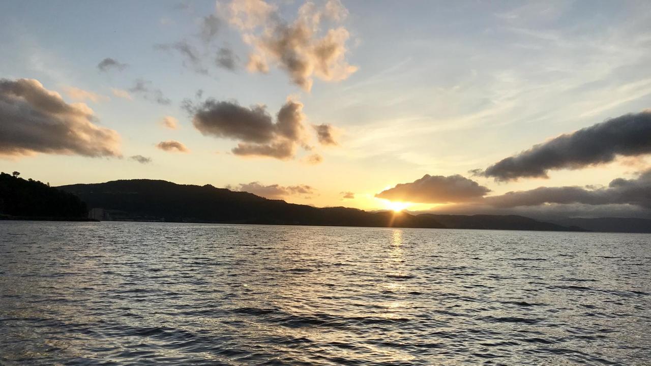 Im Vordergrund das Meer, auf dem die untergehenden Sonnenstrahlen glitzern. Im Hintergrund: eine Hügellandschaft, hinter der die Sonne gerade verschwindet. Am blauen Himmel vereinzelte langgezogene Wolken. Im Vordergrund das Meer, auf dem die untergehenden Sonnenstrahlen glitzern. Im Hintergrund: eine Hügellandschaft, hinter der die Sonne gerade verschwindet. Am blauen Himmel vereinzelte langgezogene Wolken.