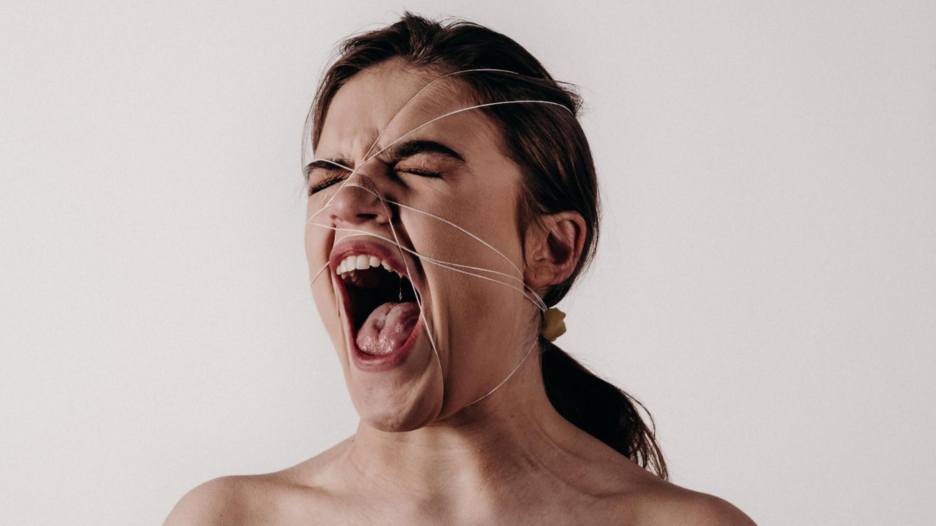 Eine Frau schreit mit weit geöffneten Mund, während ihr Gesicht von Schnüren eingesperrt wird.