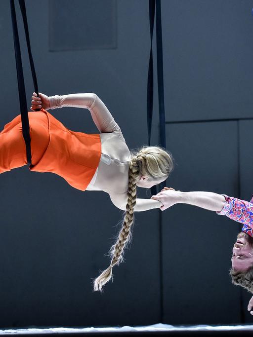 Links hängt eine Schauspielerin an einem Seil über der Bühne, sie hält rechts einen Schauspieler an der Hand, der kopfüber herunterhängt
