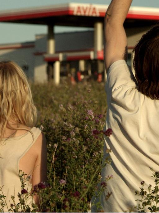 Die Zwillinge Elena (Julia Zange) und Robert (Josef Mattes) auf einer Wiese mit Blick auf eine Tankstelle im Hintergrund.