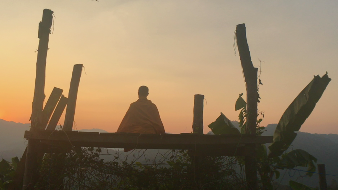 Im Still aus "Weltreise mit Buddha - Auf der Suche nach Glückseligkeit" sitzt ein buddhistischer Mönch auf einer Bank und schaut in den Sonnenuntergang.
