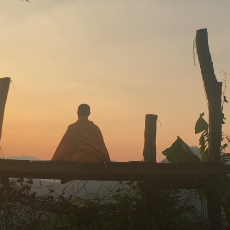 Im Still aus "Weltreise mit Buddha - Auf der Suche nach Glückseligkeit" sitzt ein buddhistischer Mönch auf einer Bank und schaut in den Sonnenuntergang.