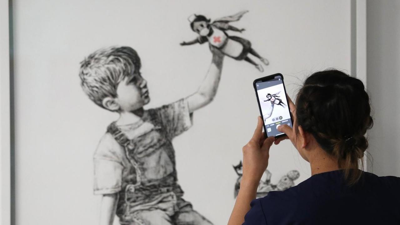 Eine Krankenschwester fotografiert mit ihrem Handy das neue Banksy-Kunstwerk: Es zeigt einen Jungen, der mit einer Krankenschwester als Superheldin spielt. Eine Krankenschwester fotografiert mit ihrem Handy das neue Banksy-Kunstwerk: Es zeigt einen Jungen, der mit einer Krankenschwester als Superheldin spielt.