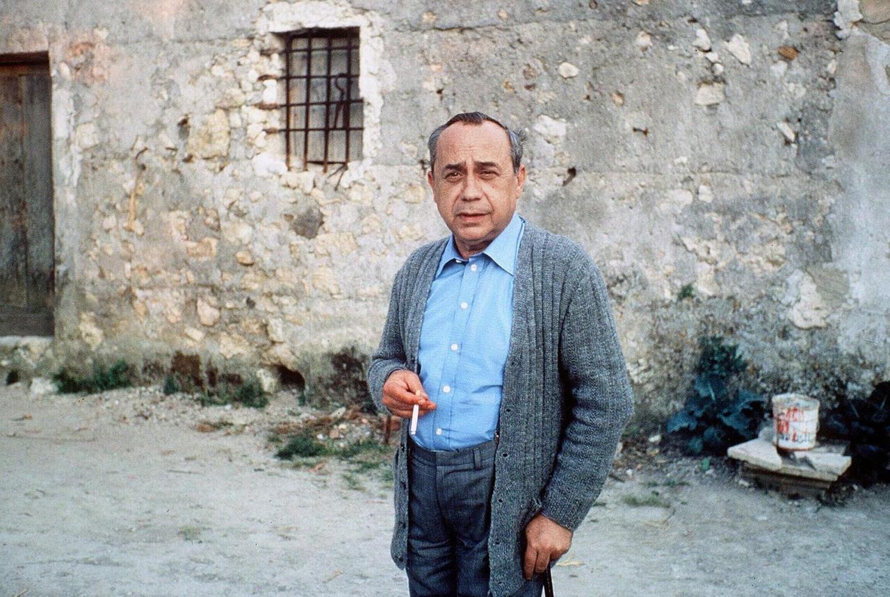 Ein Farbfoto zeigt einen Mann reiferen Alters in grauer Strickjacke mit Stock und Zigarette jeweils in in einer Hand in einem dörflich anmutenden Kontext vor einer Steinmauer.