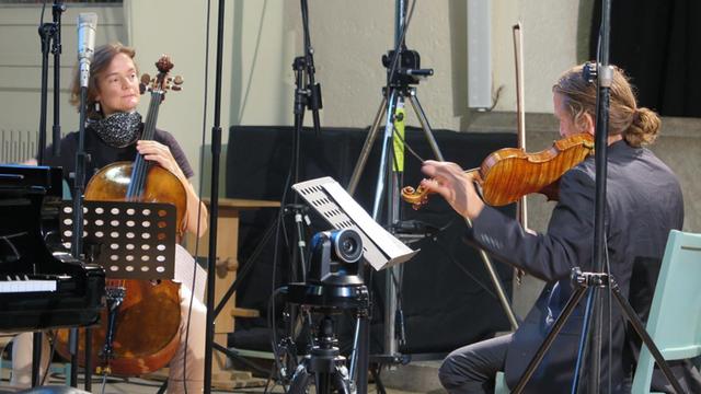 Frau mit Cello und Mann mit Geige stimmen ihre Instrumente zwischen Mikrofonständern
