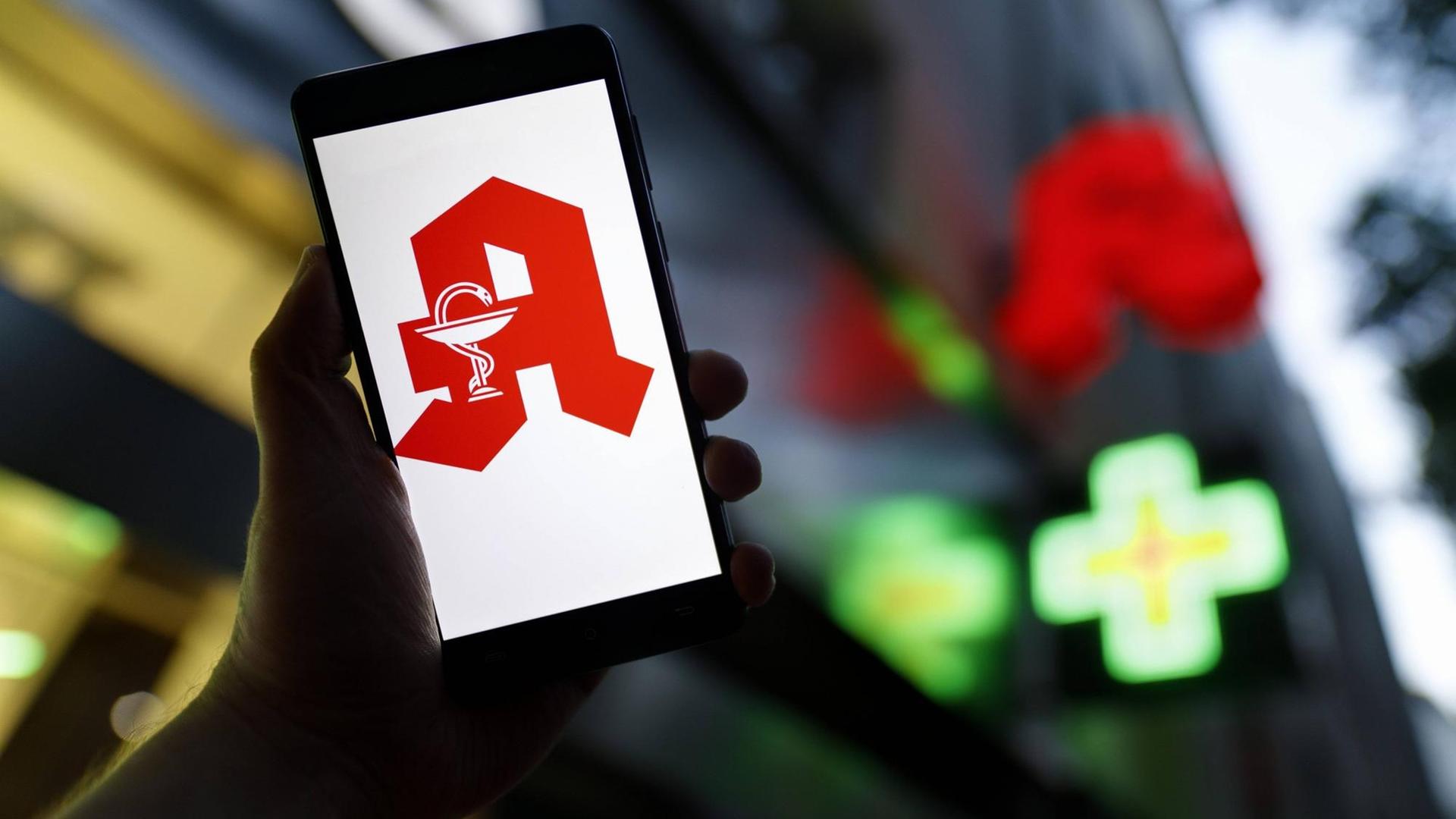 Das Apotheken-Logo erscheint auf einem Smartphone. Im Hintergrund ist eine Apotheke zu erkennen. Das Apotheken-Logo erscheint auf einem Smartphone. Im Hintergrund ist eine Apotheke zu erkennen.