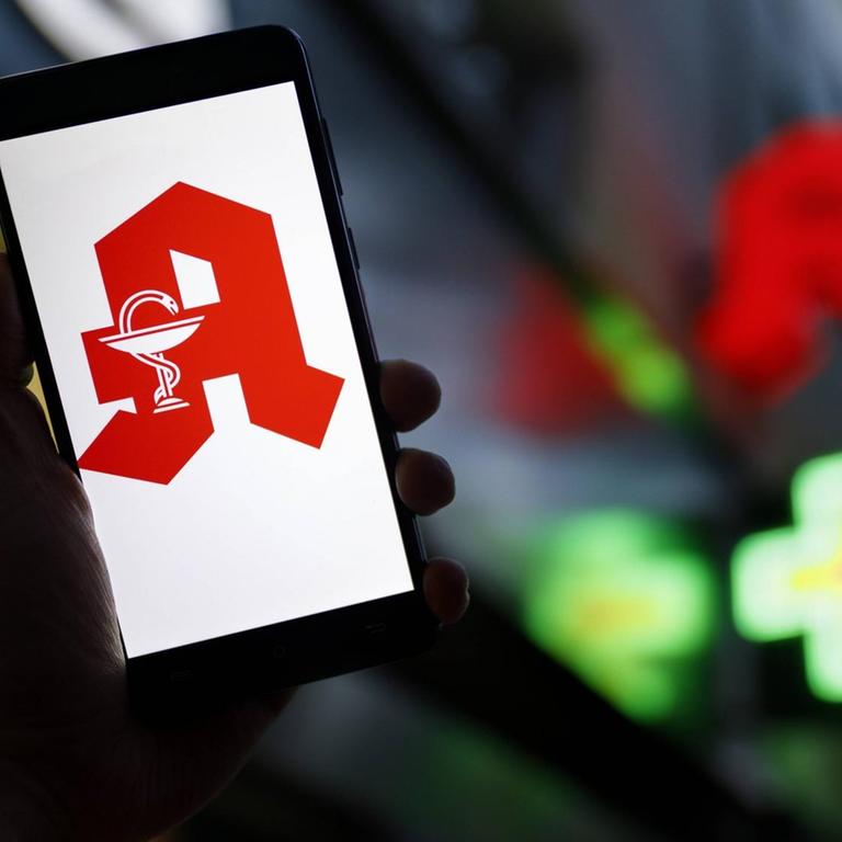 Das Apotheken-Logo erscheint auf einem Smartphone. Im Hintergrund ist eine Apotheke zu erkennen.