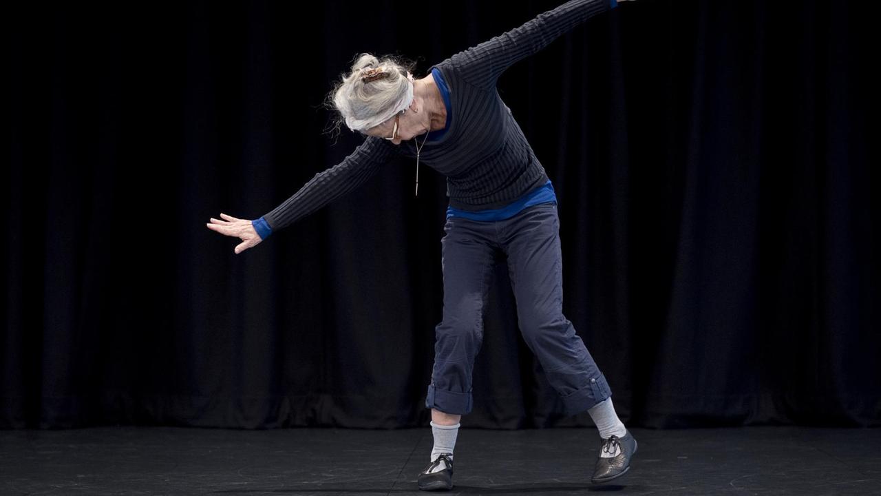 Die Choreografin Deborah Hay auf der Bühne, den Blick zum Boden gerichtet, die Arme auseinandergestreckt, den Oberkörper leicht nach unten und nach links geneigt.