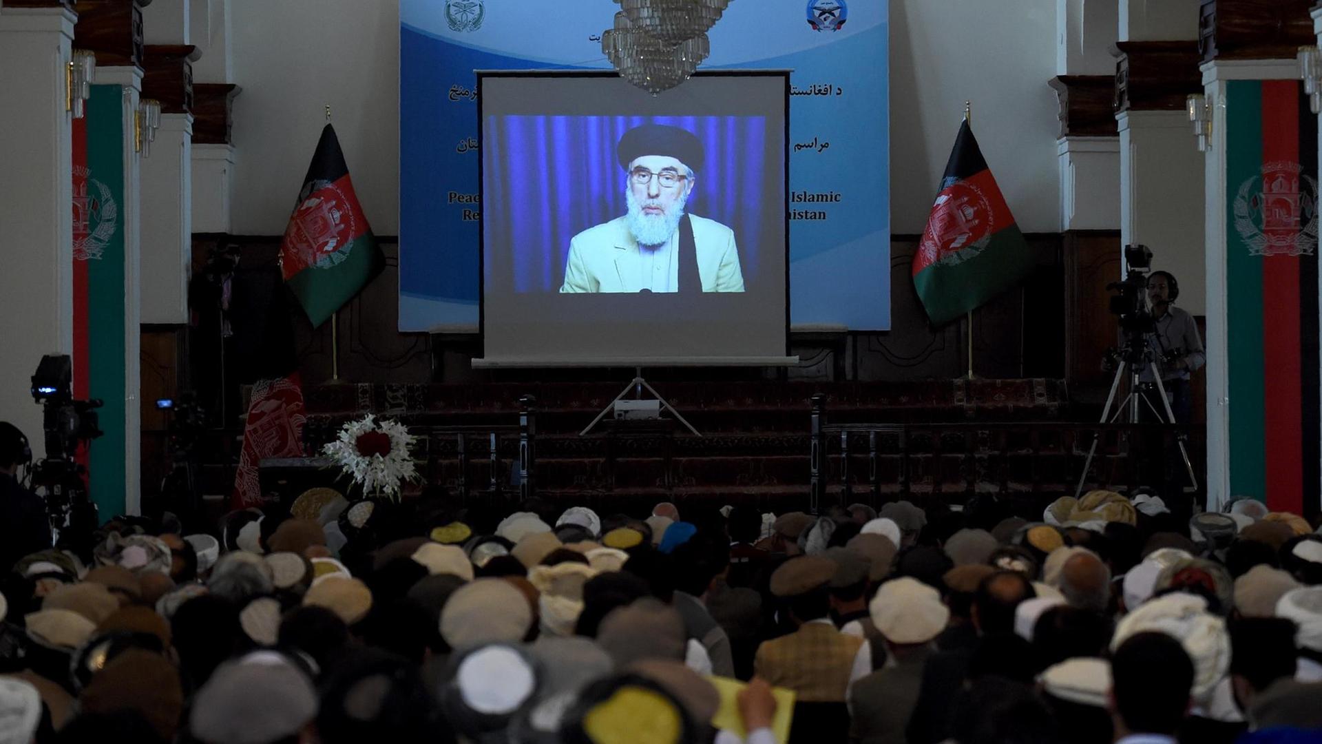 Auf einem Bildschirm in einem Saal ist der afghanische Kriegsherr Gulbuddin Hekmatyar zu sehen. Er unterschreibt gerade den Friedensvertrag mit der Regierung. Auf einem Bildschirm in einem Saal ist der afghanische Kriegsherr Gulbuddin Hekmatyar zu sehen. Er unterschreibt gerade den Friedensvertrag mit der Regierung.