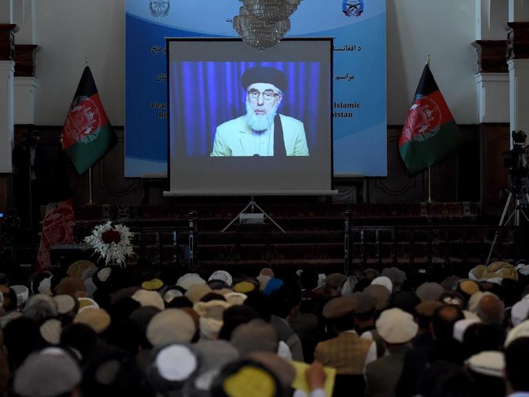 Auf einem Bildschirm in einem Saal ist der afghanische Kriegsherr Gulbuddin Hekmatyar zu sehen. Er unterschreibt gerade den Friedensvertrag mit der Regierung.