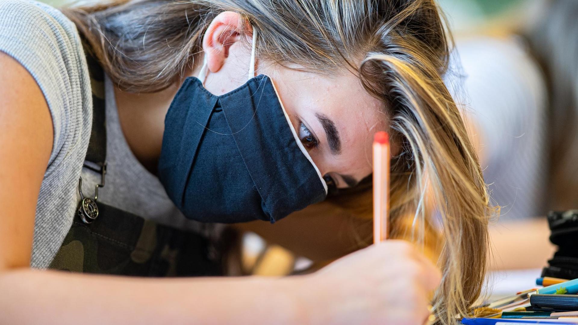 Nahaufnahme von Kopf und Schultern der Schülerin, die ein blaue Maske trägt und mit konzentriertem Blick schreibt. Nahaufnahme von Kopf und Schultern der Schülerin, die ein blaue Maske trägt und mit konzentriertem Blick schreibt.