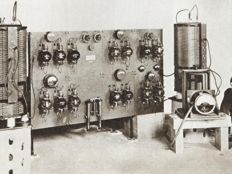 Schwarzweißaufnahme eines Radiosprechers der vor einer historischen Radioanlage sitzt.