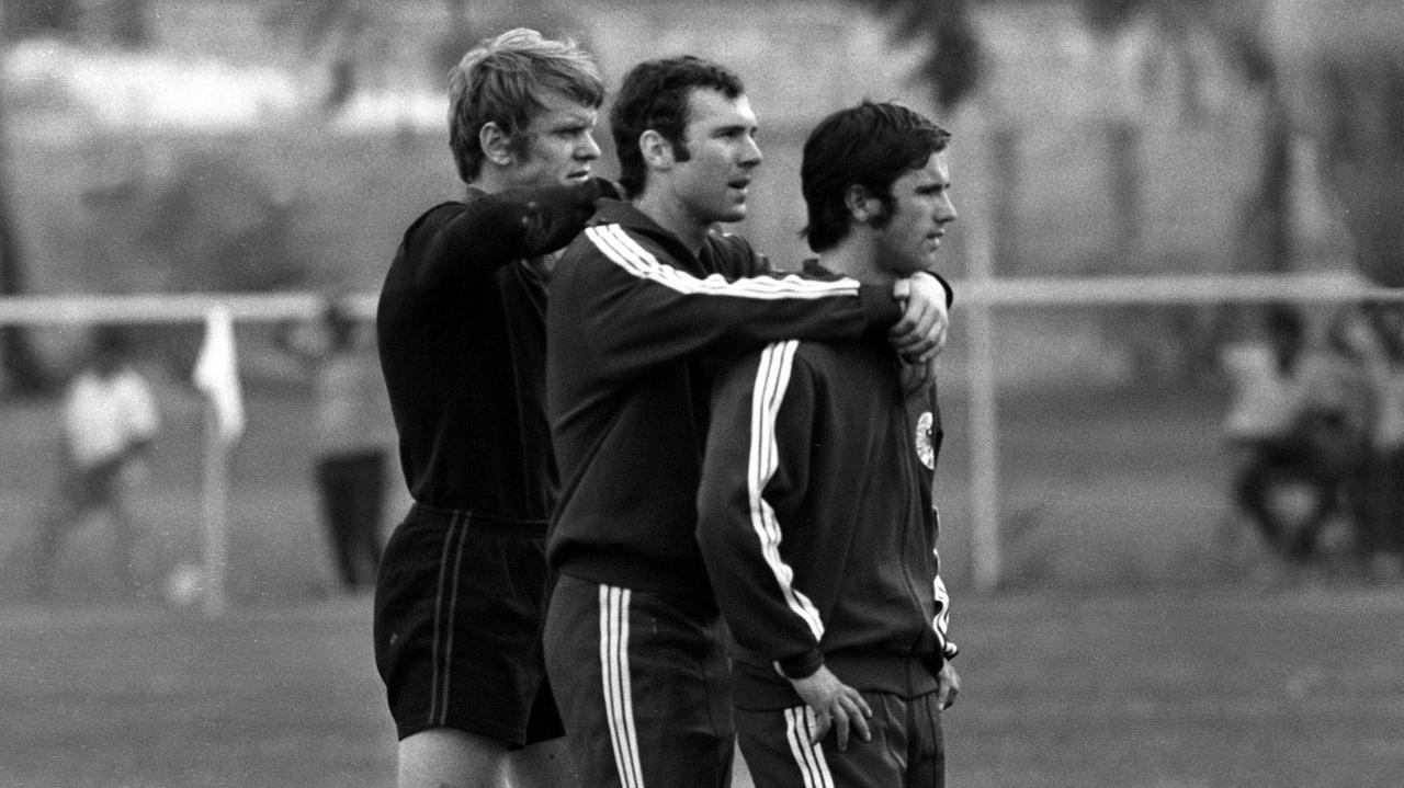 Sepp Maier, Franz Beckenbauer und Gerd Müller stehen hintereinander am Rande eines Fußballplatzes und schauen dem Training zu. Sepp Maier hat die Hände auf Beckenbauers Schultern, dieser hat die Hände auf Müllers Schultern.