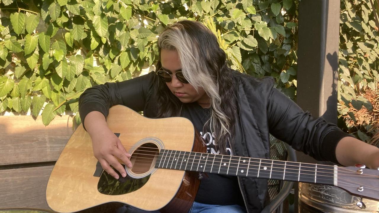 Eine junge Frau mit langen Haaren beugt sich auf einer Holzbank im Grünen über ihre Gitarre. Eine junge Frau mit langen Haaren beugt sich auf einer Holzbank im Grünen über ihre Gitarre.