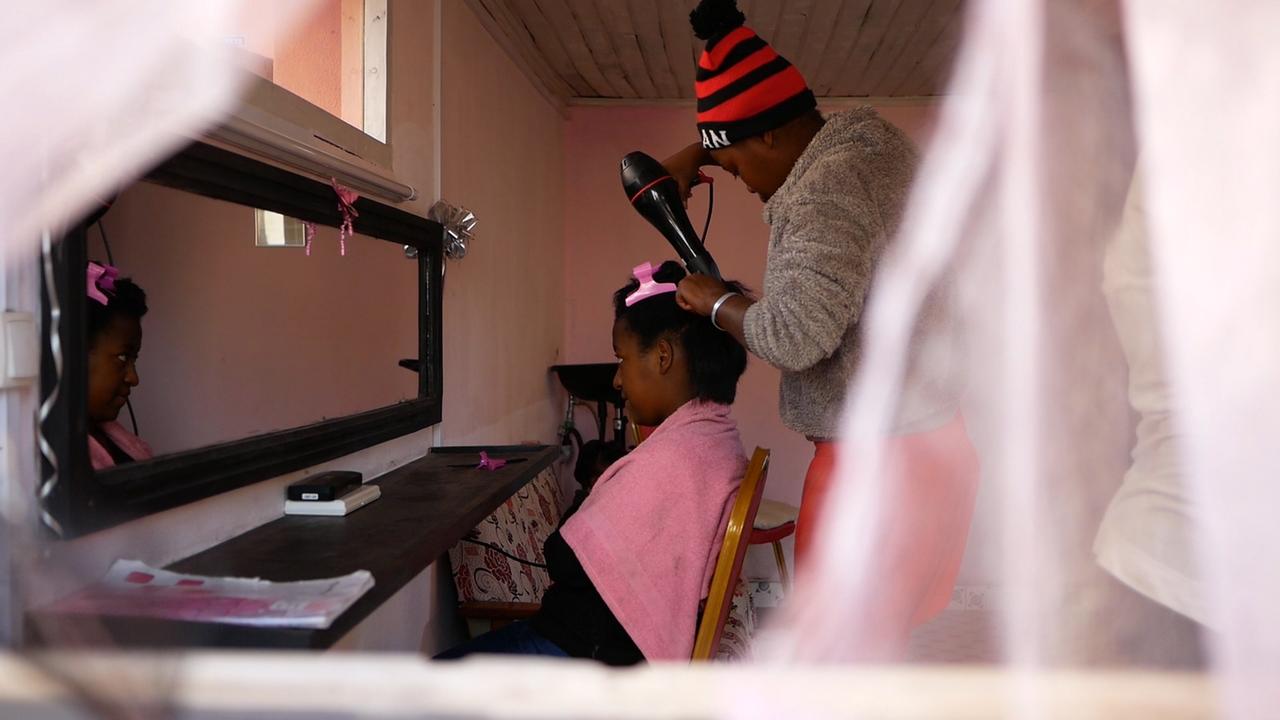 Blick durch ein Fenster in einen Friseursalon, in dem ein junges Mädchen die Haare eines weiteren jungen Mädchens fönt. Blick durch ein Fenster in einen Friseursalon, in dem ein junges Mädchen die Haare eines weiteren jungen Mädchens fönt.