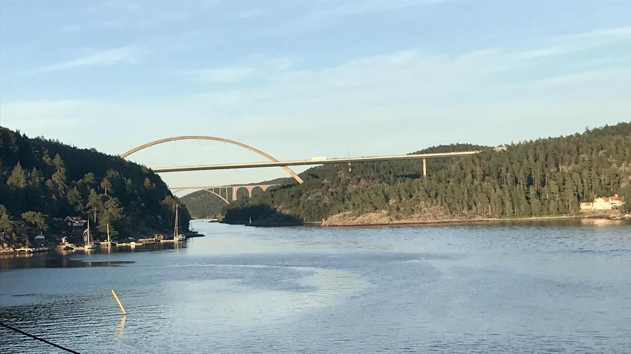Über ein breites Gewässer spannt sich hoch oben zwischen bewaldeten Bergen eine schmale helle Brücke. Über ein breites Gewässer spannt sich hoch oben zwischen bewaldeten Bergen eine schmale helle Brücke.