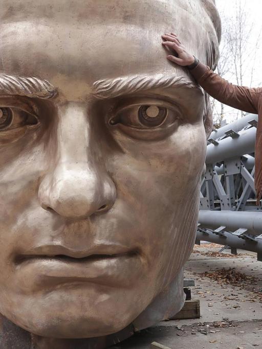 Der russische Bildhauer Andrei Korobtsov posiert in lockerer Haltung neben dem Kopf eines Denkmals. Der Kopf ist sichtlich größer als er selbst.
