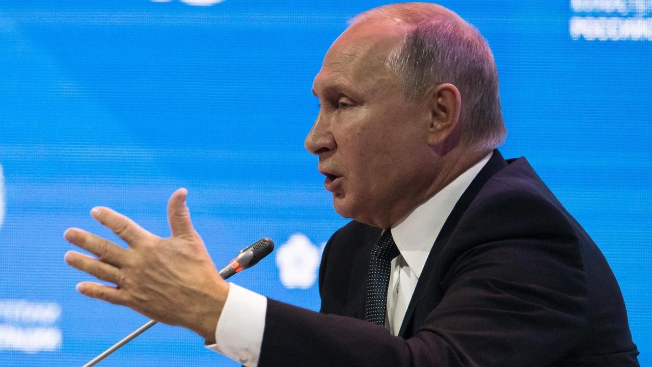Man sieht Putin redend und gestikulierend von der Seite am Mikrofon vor einer blauen Videowand.