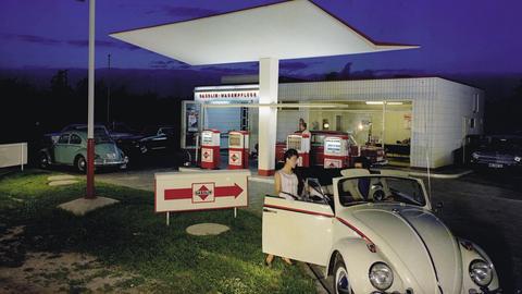 Vor einer Tankstelle steht ein beigebrauner Käfer Cabrio, auf der Beifahrerseite steigt gerade eine junge Frau ein. Aufnahme von 1958.