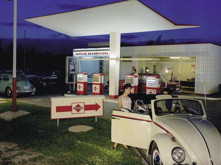 Vor einer Tankstelle steht ein beigebrauner Käfer Cabrio, auf der Beifahrerseite steigt gerade eine junge Frau ein. Aufnahme von 1958.
