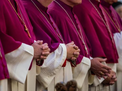 Sechs katholische Bischhöfe stehen in violetten Soutanen mit weißen Chorhemden gekleidet während einer Messe in einer Bank. Sie haben die Hände zum Gebet gefaltet.