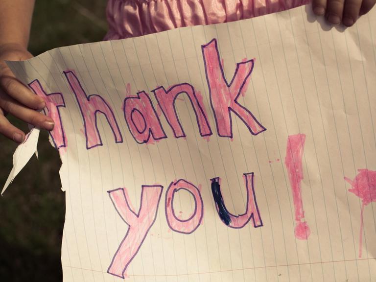 Ein kleines Mädchen in einem rosa Kleid hält ein selbstgemaltes Schild: 'thank you!'.