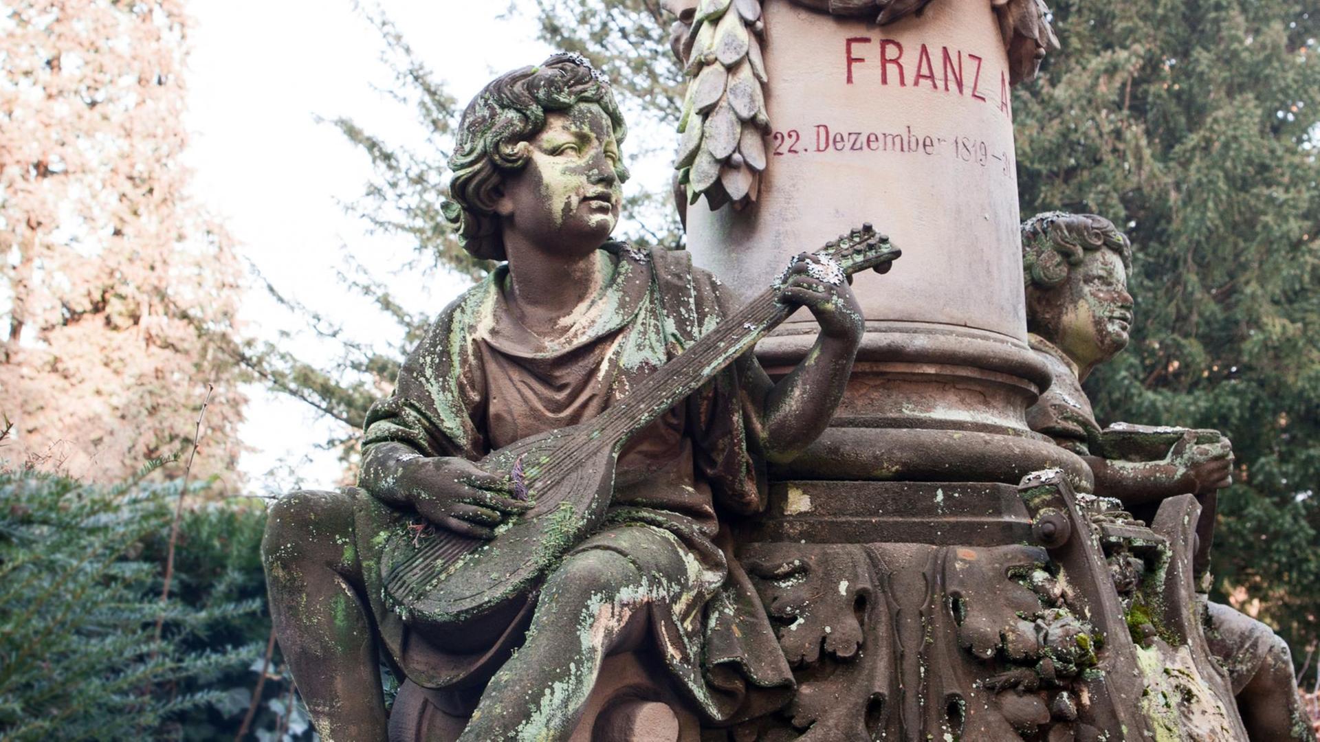 Zwei moosbewachsene Putten mit Instrumenten rahmen ein Grabmal ein. Im Hintergund stehen Nadelbäume