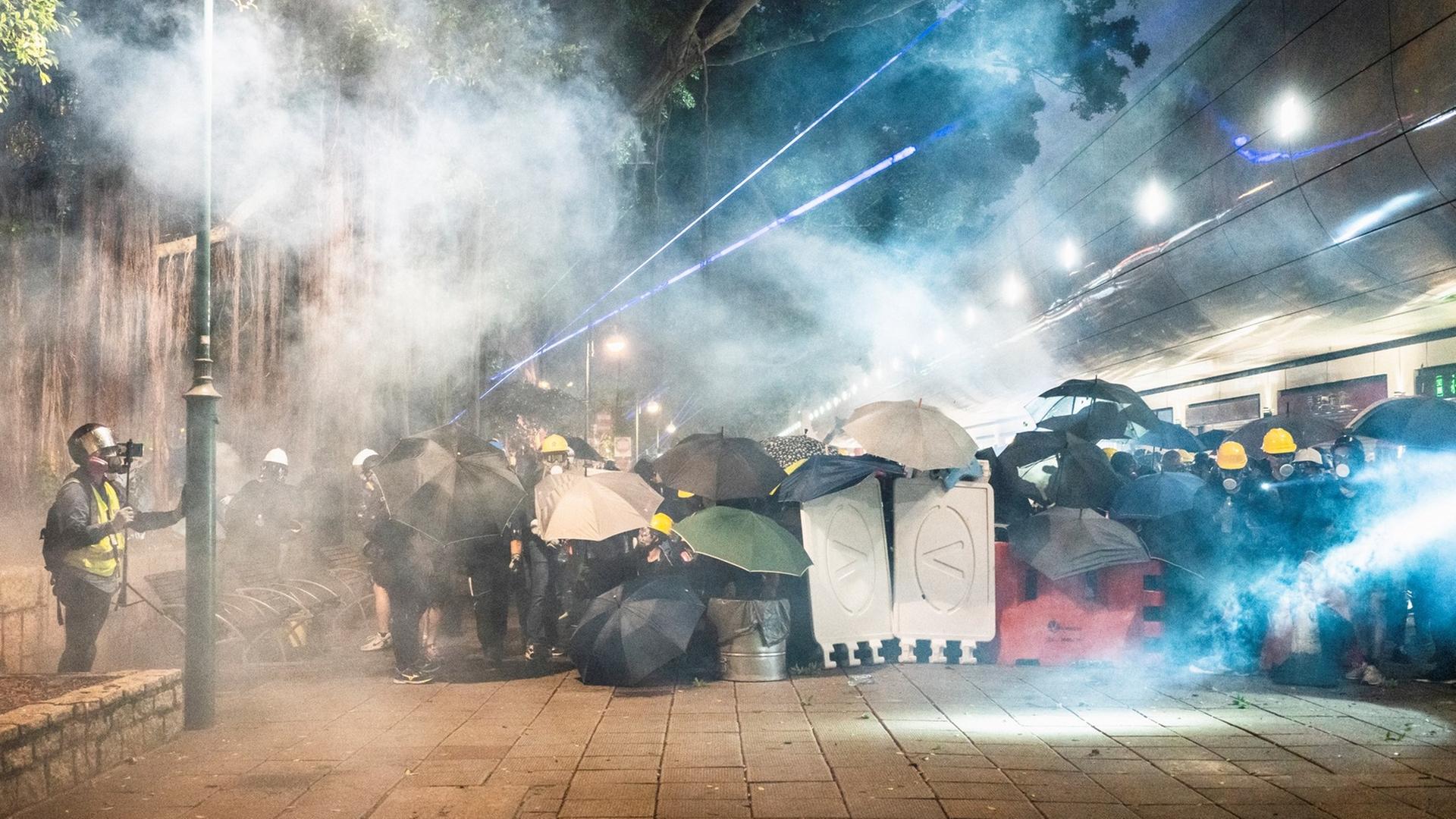 Demonstranten in Tsim Sha Tsui in Hongkong verstecken sich im Abendlicht hinter Regenschirmen und anderen Gegenständen vor dem sie umwabernden Tränengas. Demonstranten in Tsim Sha Tsui in Hongkong verstecken sich im Abendlicht hinter Regenschirmen und anderen Gegenständen vor dem sie umwabernden Tränengas.