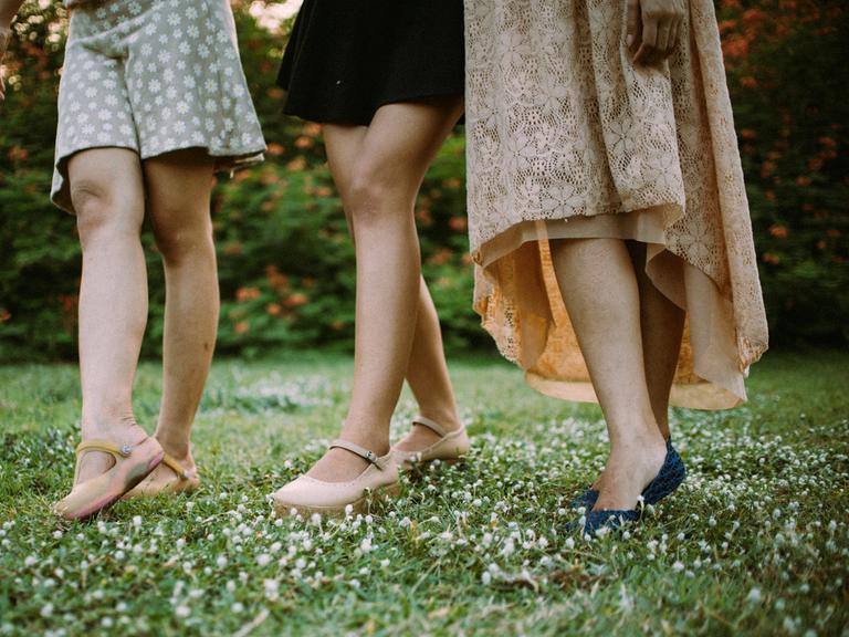 Die nackten Beine von drei Frauen in Röcken und Schuhen sind zu sehen. Sie spazieren über eine Wiese.