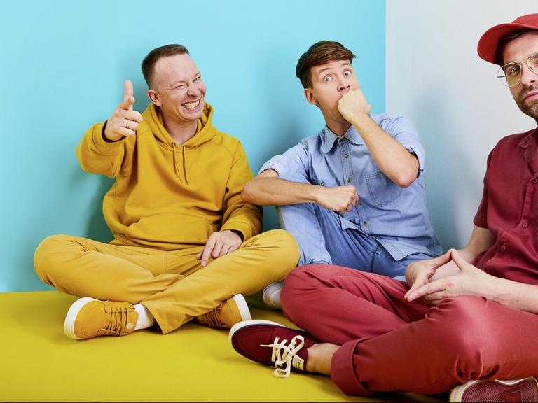 Ein Porträt der drei Bandmitglieder von "Deine Freunde" in farbenfroher Kleidung.