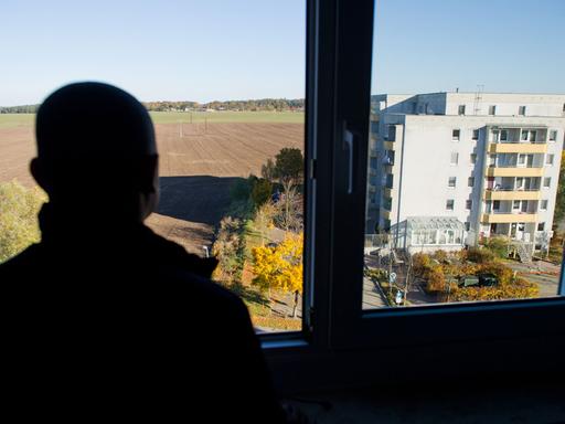 Ein Asylbewerber blickt am 26.10.2012 aus einem Fenster eines Asylbewerberheims in Wolgast in Mecklenburg-Vorpommern. Vor dem Fenster ist ein Plattenbau und flache Landschaft mit Äckern, Wiesen und Wald zu sehen.
