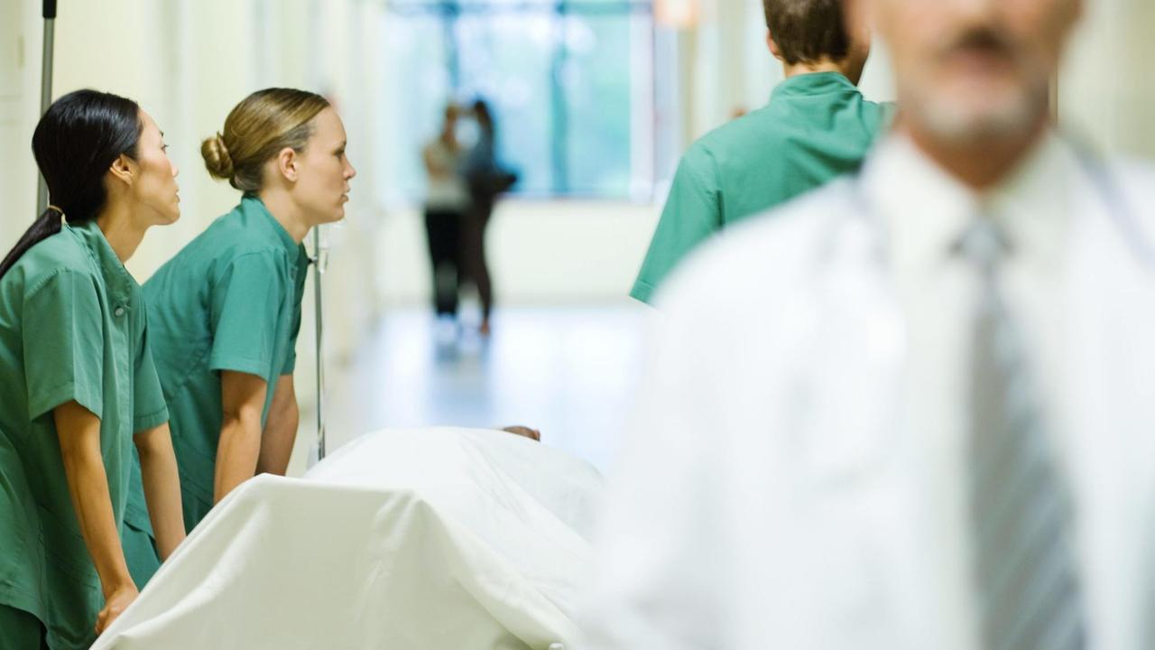 Junge Ärzte schieben eine Krankenhausliege durch den Gang, ein Arzt läuft im Vordergrund durch das Bild. Junge Ärzte schieben eine Krankenhausliege durch den Gang, ein Arzt läuft im Vordergrund durch das Bild.