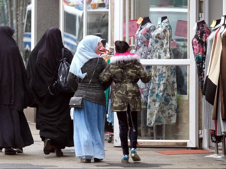 Zwei in schwarz verhüllte ältere Frauen und zwei jüngere Mädchen, davon eins mit mit Kopftuch, passieren ein muslimisches Bekleidungsgeschäft mit Schaufensterpuppen in Duisburg-Marxloh. 