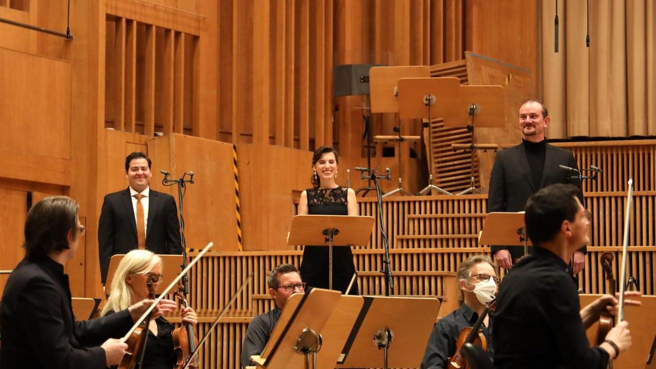 Drei Sänger lächeln stehend, da das Orchester, das vor ihnen sitzt, ihnen Beifall mit den Bögen zollt. Drei Sänger lächeln stehend, da das Orchester, das vor ihnen sitzt, ihnen Beifall mit den Bögen zollt.