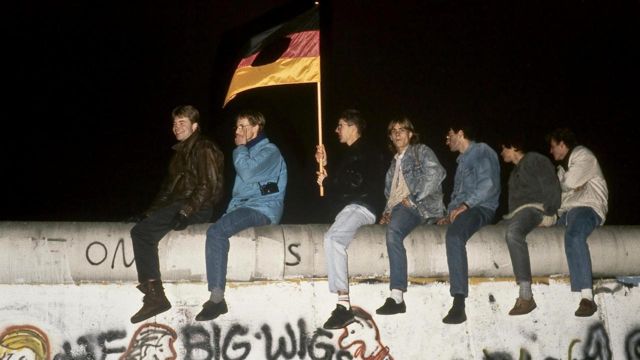Ein Gruppe junger Männer sitzt mit einer zerschnittenen DDR-Fahne auf der Mauer am Brandenburger Tor in Berlin. Ein Gruppe junger Männer sitzt mit einer zerschnittenen DDR-Fahne auf der Mauer am Brandenburger Tor in Berlin.