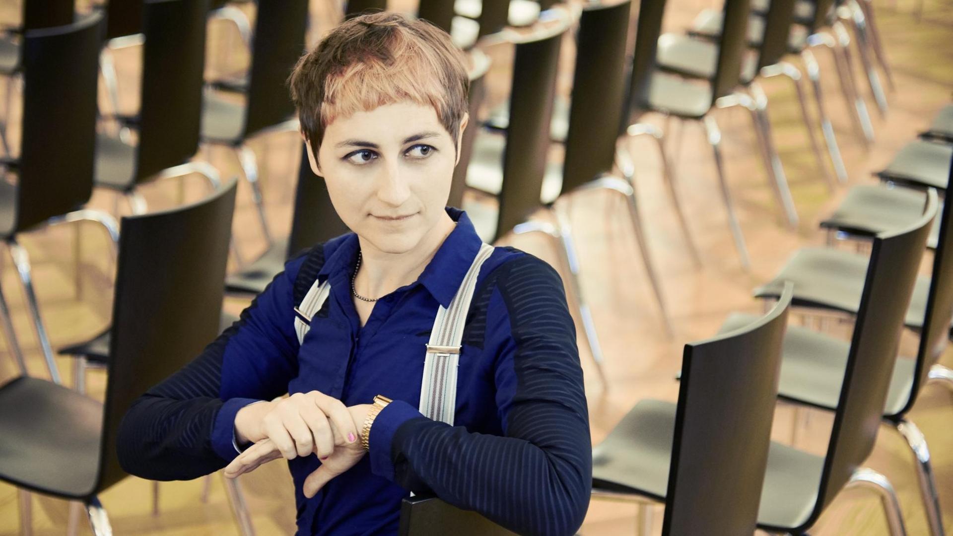 Ulrike Almut Sandig sitzt in einem Raum in einer von vielen Stuhlreihen. Die Schriftstellerin und Lyrikerin trägt ein blau-schwarzes Oberteil an, die Haare trägt sie kurz. Ulrike Almut Sandig sitzt in einem Raum in einer von vielen Stuhlreihen. Die Schriftstellerin und Lyrikerin trägt ein blau-schwarzes Oberteil an, die Haare trägt sie kurz.