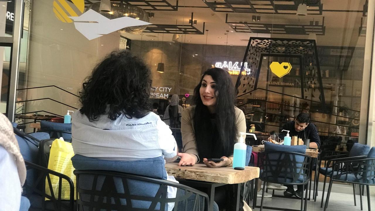 In einem Café sitzt eine junge Frau mit vollem schwarzem langem Haar gegenüber von einem Mann an einem Tisch.