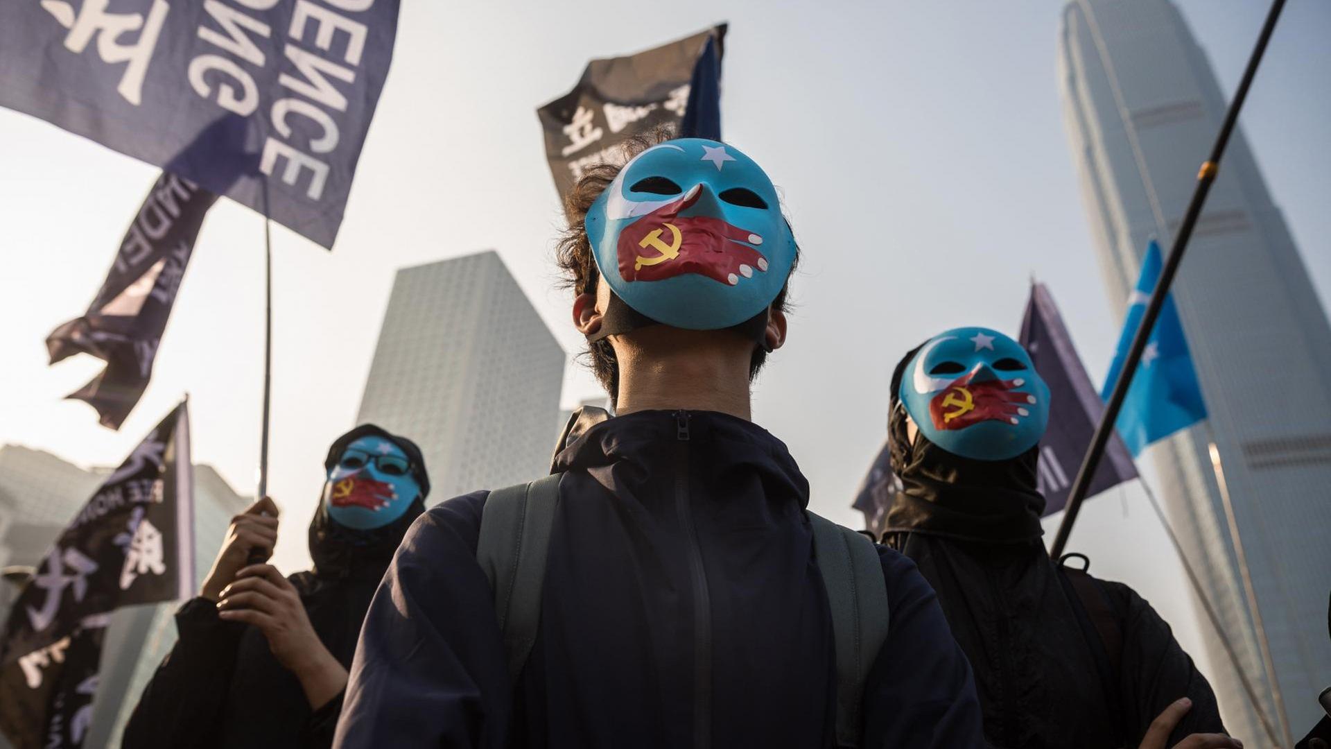 Schwarzgekleidete Demonstranten tragen blaue Gesichtsmasken. Darauf gemalt sind Hände mit einer chinesischen Flagge in Form einer Hand, die den Mund zuhält. Schwarzgekleidete Demonstranten tragen blaue Gesichtsmasken. Darauf gemalt sind Hände mit einer chinesischen Flagge in Form einer Hand, die den Mund zuhält.