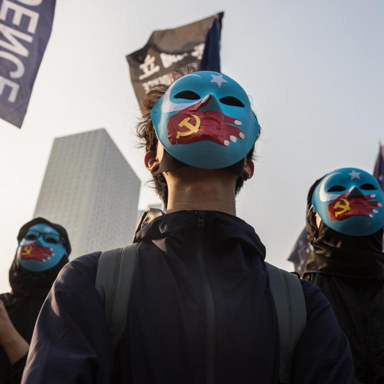 Schwarzgekleidete Demonstranten tragen blaue Gesichtsmasken. Darauf gemalt sind Hände mit einer chinesischen Flagge in Form einer Hand, die den Mund zuhält.