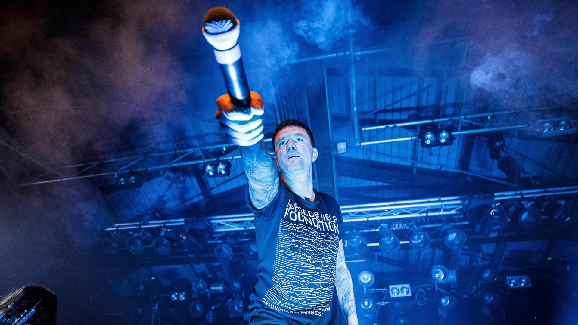 Marcus Bischoff, Sänger der Band "Heaven Shall Burn" auf der Bühne, mit einem Mikrofon in der Hand, das er in Richtung des Publikums hält. Marcus Bischoff, Sänger der Band "Heaven Shall Burn" auf der Bühne, mit einem Mikrofon in der Hand, das er in Richtung des Publikums hält.
