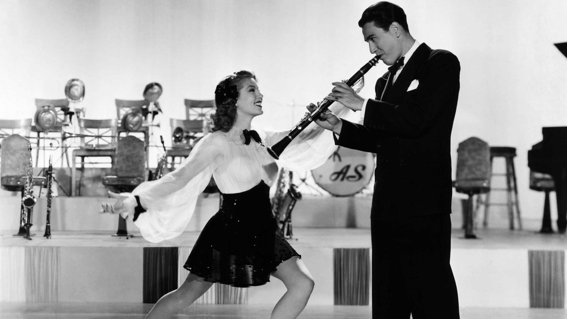 Schwarzweiß-Standbild eines alten Films, auf dem eine Frau in tanzender Haltung einen Mann anstrahlt, der in schwarzem Anzug, Klarinette spielt. Sie trägt eine weiße Bluse und einen schwarzen Rock, der bis knapp zu den Knien reicht. Im Hintergrund sind, Stühle, Saxofone und Stehlampen verschwommen zu erkennen.
