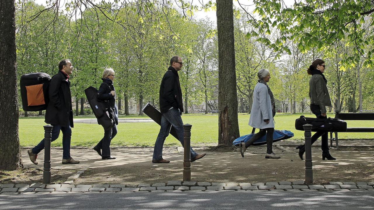 Fünf Musiker laufen mit Gepäck hintereinander durch einen Park.