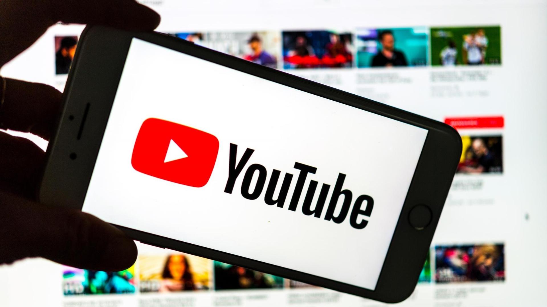 Das Logo des Video-Portals YouTube wird auf dem Display eines Smartphones angezeigt. Im Hintergrund ist auf einem Bildschirm die YouTube Homepage zu sehen. Das Logo des Video-Portals YouTube wird auf dem Display eines Smartphones angezeigt. Im Hintergrund ist auf einem Bildschirm die YouTube Homepage zu sehen.