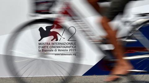 Vor dem offiziellen Logo der 72. Filmfetspiele in Venedig 2015 fährt ein Mann Fahrrad. Schemenhaft sind die Beine und das Fahrrad zu sehen.